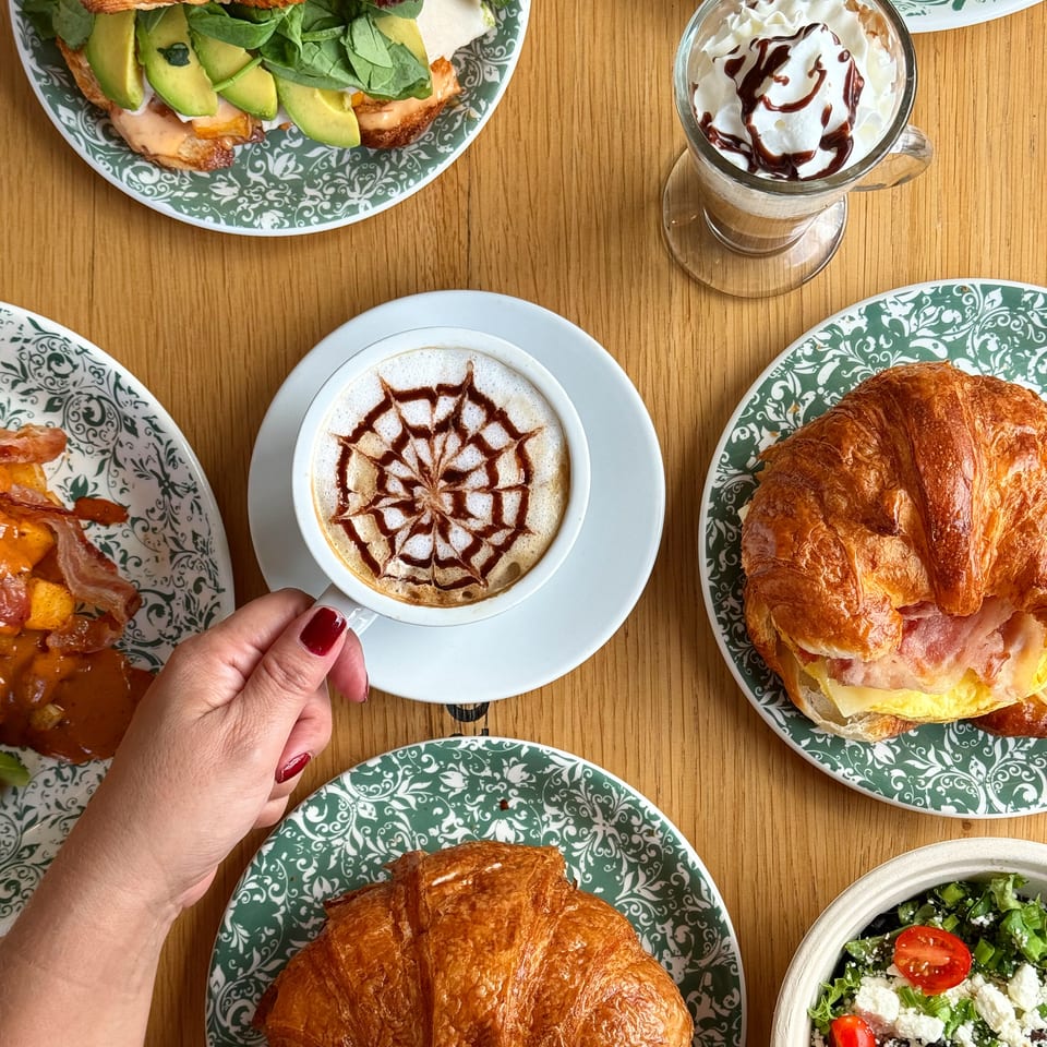 Overhead spread of croissant sandwiches, salads, chocolate-topped coffee, whipped cream dessert, and roasted potatoes on a wooden table.