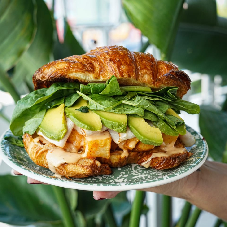 Loaded croissant sandwich with avocado, spinach, roasted potatoes, and creamy sauce on a patterned plate amid tropical greenery.