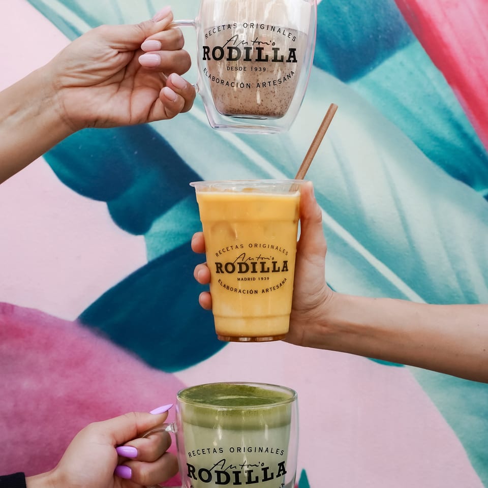 Hands hold three Antonio Rodilla drinks—coffee, orange juice, and a green beverage—against a colorful abstract background.