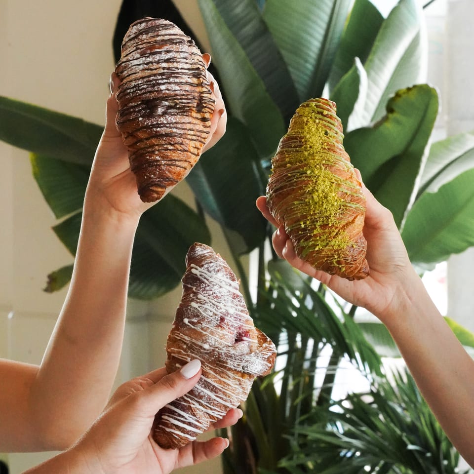 Hands hold chocolate-drizzled and pistachio-topped croissants against a backdrop of lush indoor plants.