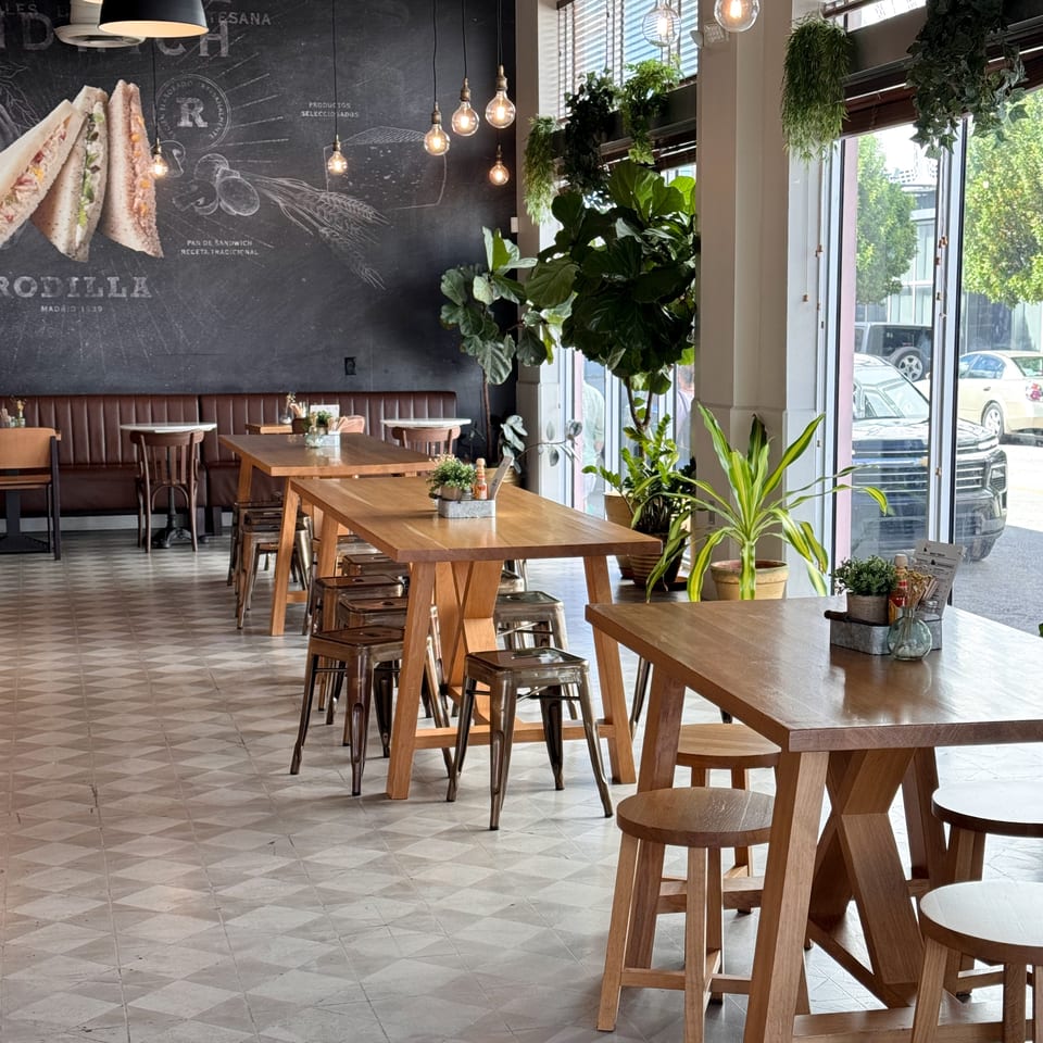 Bright restaurant dining room with wooden tables, stools, pendant lights, patterned tile flooring, lush plants, and sandwich mural.