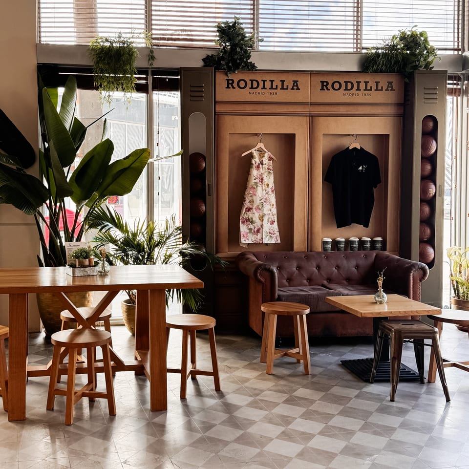Rodilla Madrid 1939 restaurant interior with plants, wooden tables, leather sofa, merchandise displays, and patterned tile flooring.